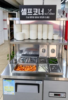 반찬 셀프코너 — «셀프코너 SELF CORNER 드실 양만큼 담아서 맛있게 드세요» 판 아래 쌓인 그릇과 깍두기·양파·풋고추 통