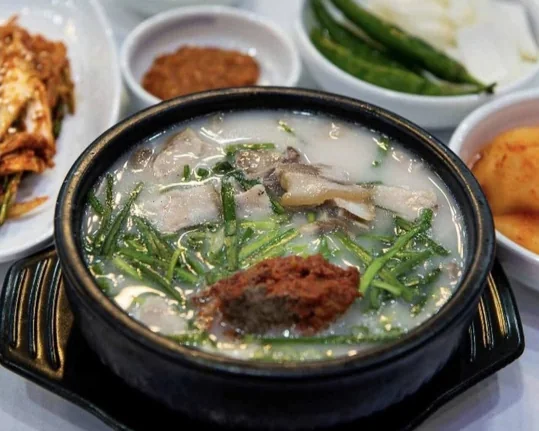 뚝배기 국밥 가까이 — 뽀얀 국물 속 고기, 위에 부추와 붉은 다대기, 옆으로 깍두기·풋고추와 양파·장 그릇