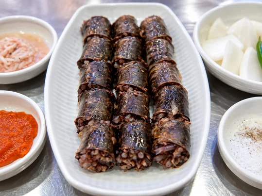 병천순대 한 접시 — 곡물과 채소 소가 보이는 굵은 순대 썬 것, 둘레에 새우젓·붉은 양념장·양파와 풋고추·소금 후추 종지