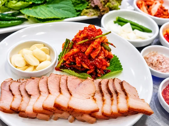 수육 한 접시 — 껍질째 도톰하게 썬 수육, 깻잎 위 붉은 무침과 부추, 마늘 종지, 뒤로 쌈채소·풋고추·새우젓