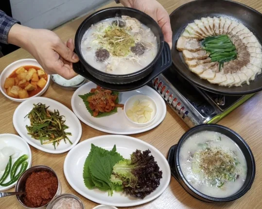 수육과 국밥 한 상 — 두 손으로 든 순대 국밥 뚝배기, 버너 위 팬에 수육과 부추, 쌈채소·마늘·깍두기·부추무침 같은 반찬